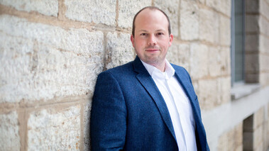 Porträt von Sebastian Klembt in blauem Sakko vor einer Sandsteinwand, leicht lächelnd.  Dateiname: reinraumlabor-video-screenshot-20210226.jpg